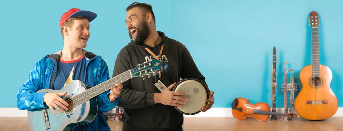 A male participant joyfully holds a guitar while a TLC support worker holds a small drum, creating a harmonious and happy musical moment in our engaging day programs.