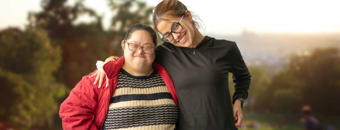 A female TLC support worker stands alongside a happy female participant, with her arm around the participant's shoulder, as they both look at the camera, exemplifying the inclusive and supportive environment of our multilingual services.