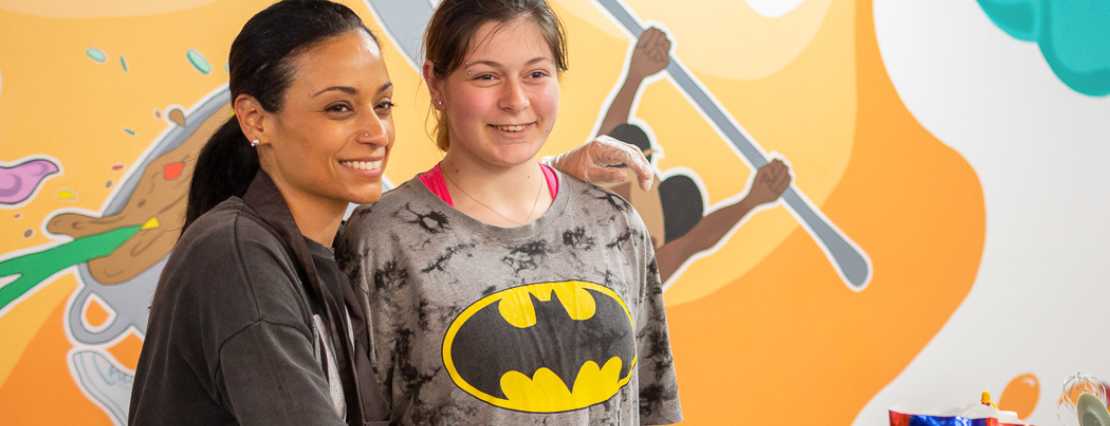 A cheerful support worker shares a smile with a participant dressed in a Batman garment, adding fun and excitement to one of our engaging day programs.